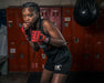 Person in boxing stance with red gloves and black shorts in front of red lockers.
