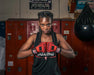 Boxer in a gym with red gloves and black and red outfit, standing in front of red lockers.