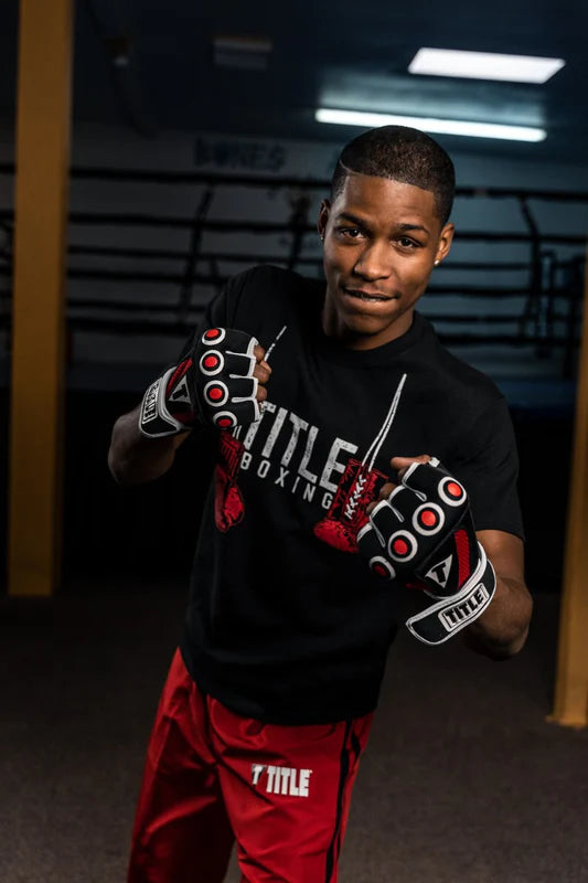 A man in a boxing stance wearing a black T-Title Boxing t-shirt, red boxing shorts, and black and red boxing gloves in a gym setting.